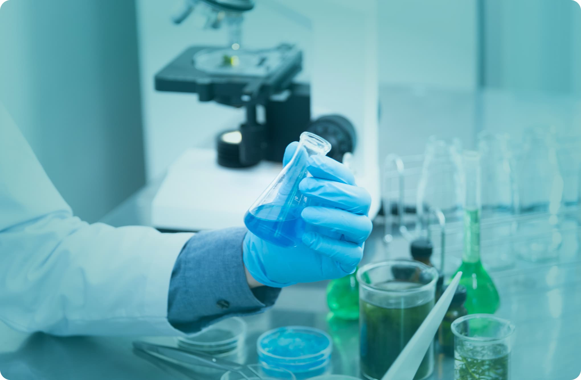 Scientist wearing gloves holding a flask with blue liquid in a laboratory, with a microscope and glassware in the background.